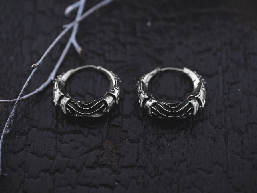 A pair of Kaya Movement’s Bio Organic Silver Hoop Earrings, featuring futuristic, Alien Xenomorph-inspired designs by HR Giger, are placed on a textured dark surface. Thin white branches lie across the background, providing contrast against the rich black backdrop. The blend of classic motifs and cyberpunk style creates a striking visual for these statement men's earrings.