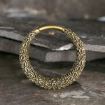 A close-up of Kaya Movement's PAWA Tribal Thick Hoop Ear Weights in Gold with Clicker, showcasing their intricate floral engravings. The ear weights are placed on a textured stone surface, which provides a rustic backdrop, enhancing their detailed design and craftsmanship at 6 gauge.