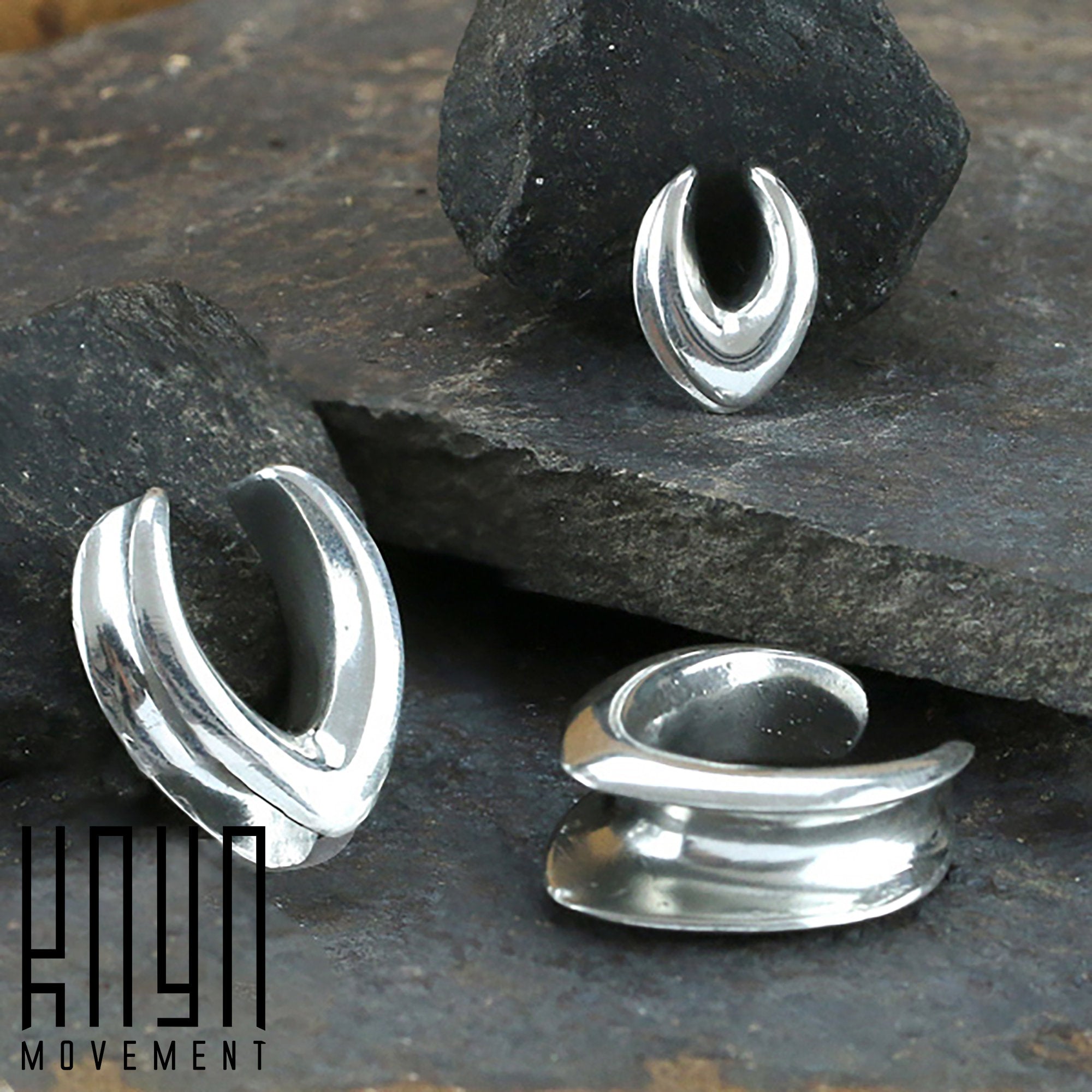 Three silver MYRA Minimalist Saddle Tunnels are displayed on a rocky surface. Each tunnel features a unique, modern design with smooth, curved shapes, resembling a subtle wishbone shape. The brand logo "Kaya Movement" appears in the bottom left corner.