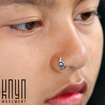 Close-up of a person's face showcasing a jeweled nose ring with a small turquoise gemstone and bead accents. The background is blurred, and the person’s mouth is slightly open. The logo “Kaya Movement” is visible in the bottom left corner, highlighting this exquisite 925 silver body jewelry piece, the SHA Beaded Flower Nose Ring in Silver & Turquoise | 20 gauge.