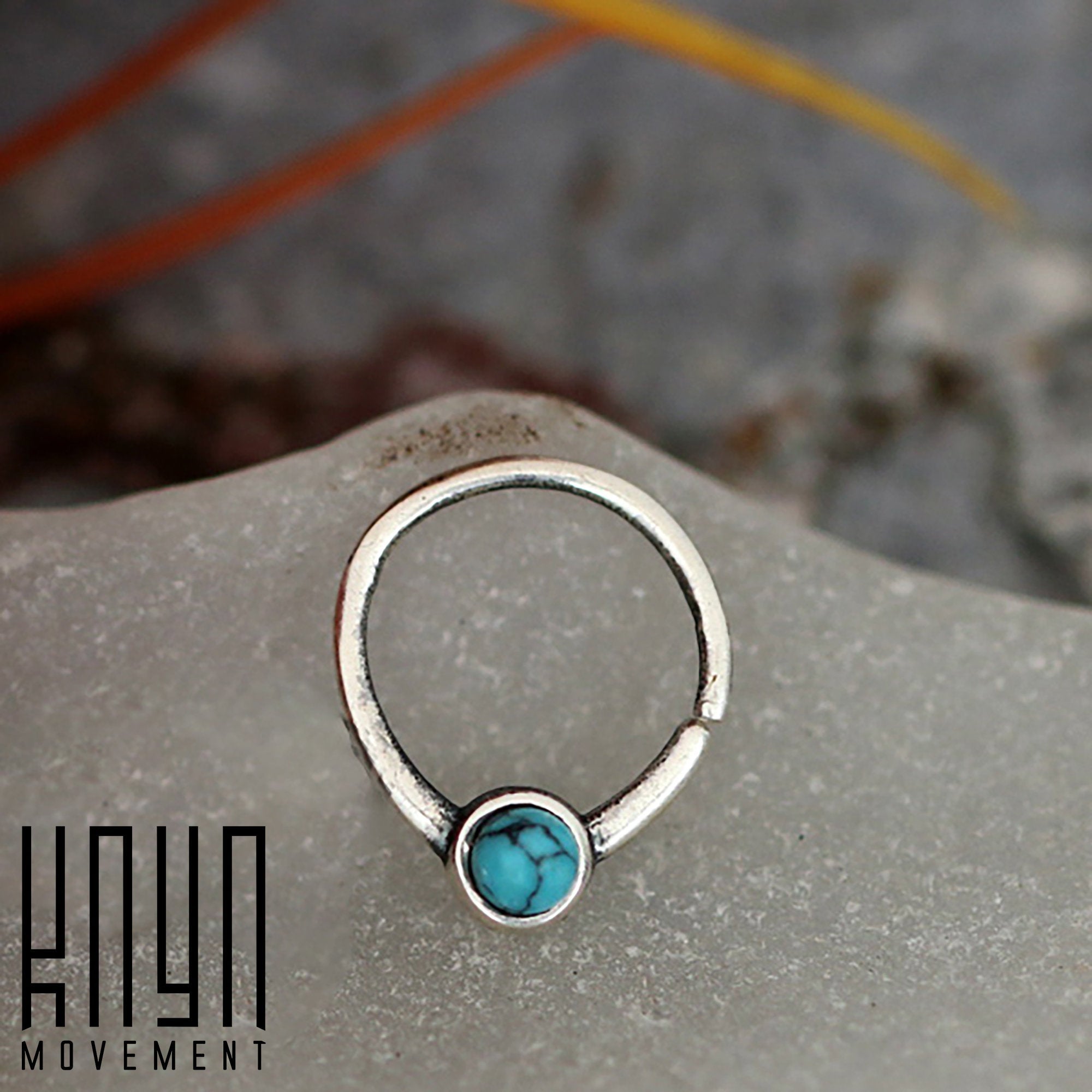 A silver NAE Seamless Septum Ring in 18 gauge, featuring a natural blue Turquoise gemstone centerpiece from Kaya Movement, is displayed on a frosted glass surface. The text "Kaya Movement" is printed in black on the bottom left corner of the image. The background is blurred, showcasing hints of grey and orange tones.