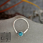 A silver NAE Seamless Septum Ring in 18 gauge, featuring a natural blue Turquoise gemstone centerpiece from Kaya Movement, is displayed on a frosted glass surface. The text "Kaya Movement" is printed in black on the bottom left corner of the image. The background is blurred, showcasing hints of grey and orange tones.
