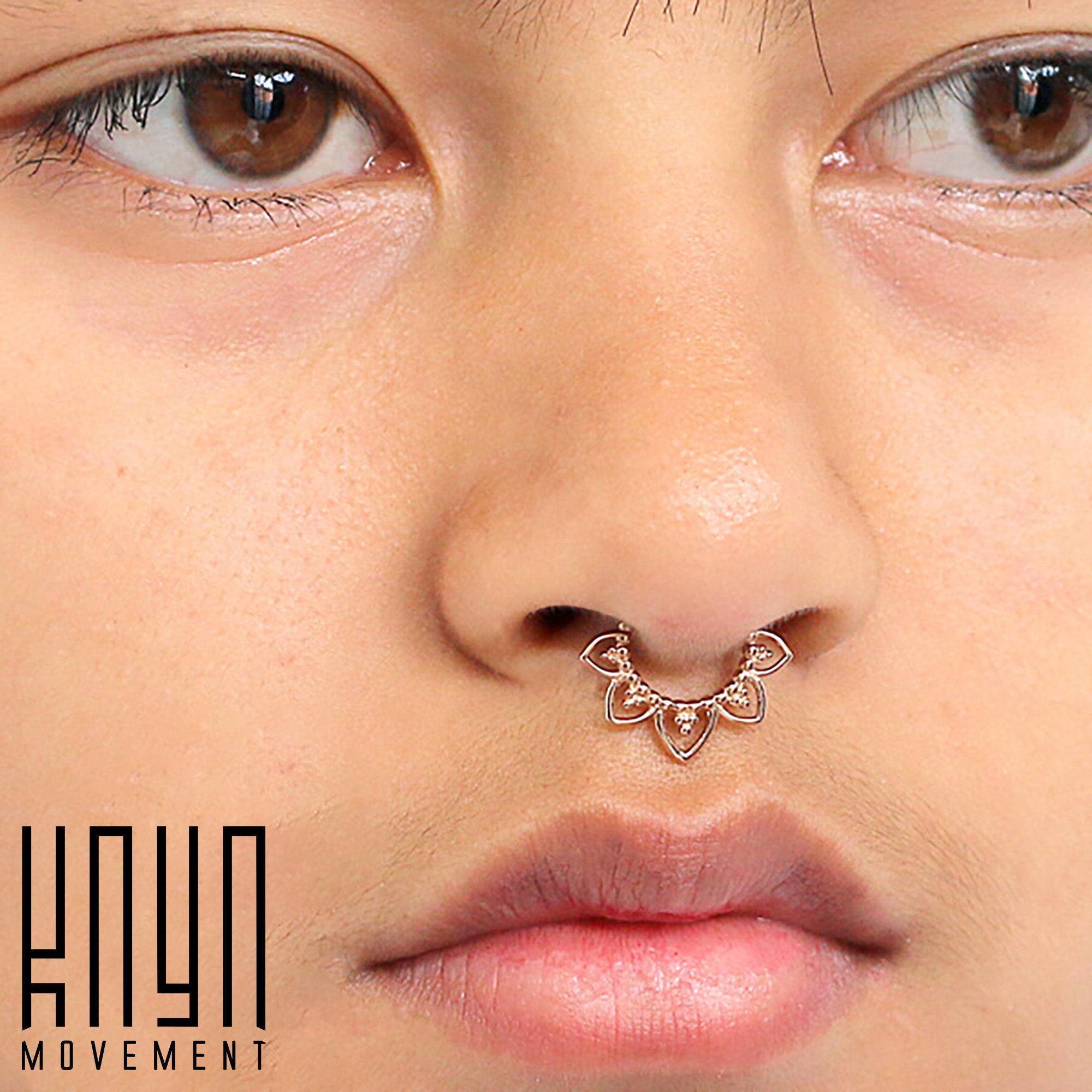 Close-up of a person's face, focusing on the MAWAR Lotus Clicker Septum Ring in Rose Gold (16 gauge) by Kaya Movement, a decorative piercing with a floral design. The brown-eyed individual has a neutral expression. The logo "Kaya Movement" is visible in the bottom left corner.