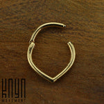 A small piece of gold body jewelry, known as the TIKA Teardrop Septum Clicker Ring in Gold (16 gauge) by Kaya Movement, featuring a minimalist design with an open, unjoined section, rests elegantly on a textured wooden surface. The ring, shaped like a teardrop or an inverted heart, subtly blends the allure of surgical steel. A logo with the text "Kaya Movement" is visible in the bottom left corner.