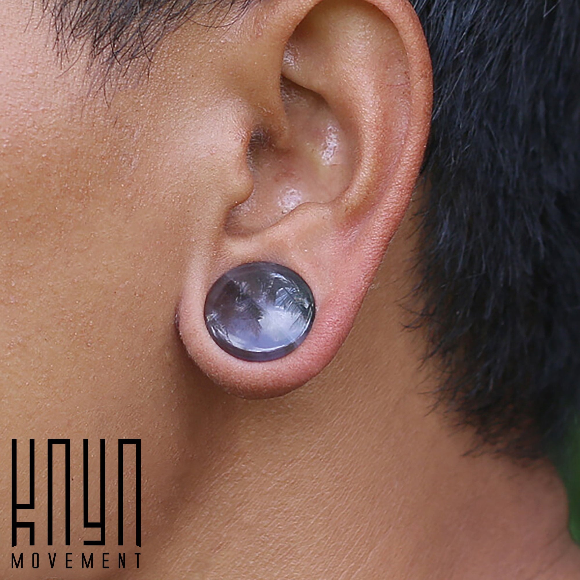 Close-up of a person's ear showcasing a black gauge earring and a glossy, round plug adorned with an amethyst stone from the Amethyst Round Stone Saddle Plugs | 4mm to 25mm gauge collection. The Kaya Movement logo is seen in the lower left corner of the image.