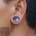 Close-up of a person's ear showcasing a black gauge earring and a glossy, round plug adorned with an amethyst stone from the Amethyst Round Stone Saddle Plugs | 4mm to 25mm gauge collection. The Kaya Movement logo is seen in the lower left corner of the image.