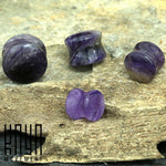 Four Amethyst Round Stone Saddle Plugs lie on a textured stone surface. Each plug is carved into a curved, unique shape, showcasing various shades of purple with subtle white streaks. In the bottom left corner, the "Kaya Movement" logo is visible in black.