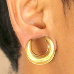 A close-up of an ear wearing a large, gold Minimalist Horseshoe Saddle Tunnel from Kaya Movement. The earlobe is stretched to accommodate the thick, shiny piece of jewelry, available in sizes ranging from 4mm to 30mm gauge. The background is neutral, putting the focus on the ear and earring.