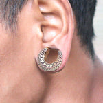 Close-up of an ear adorned with a crescent-shaped DAG Tribal Horseshoe Saddle Tunnel in Gold by Kaya Movement. The earring features intricate engravings and small, circular indentations. The person's skin tone is visible, and short, dark hair frames the ear.