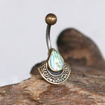 Close-up of the IKOSA Fan Belly Piercing Ring in Gold & Abalone Shell by Kaya Movement. The 14-gauge belly jewelry features a curved barbell design made from surgical steel, highlighted by a top gold-colored bead and an ornate base set with a tear-drop-shaped abalone shell. The ring is displayed against a natural wood background.