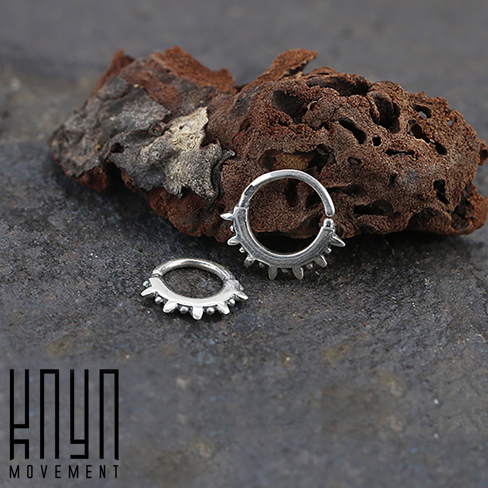 A 16-gauge SPIKE Punk Spike Nose Ring in silver from Kaya Movement is placed on a dark, textured surface with a piece of rugged rock in the background. The tiny hoop ring features small spikes around the perimeter. The text "Kaya Movement" is seen in the bottom left corner of the image.