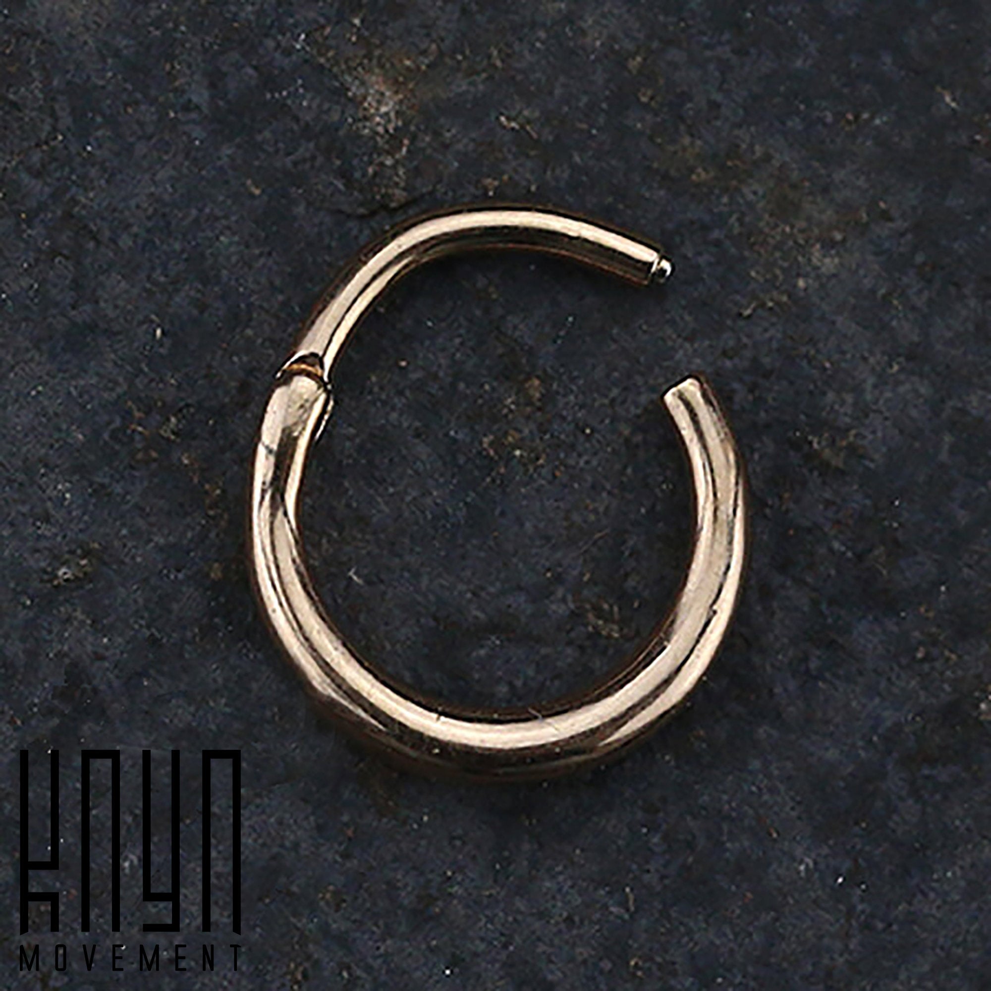 A close-up of a small, circular rose gold AMAO Clicker Nose Ring in 316L Surgical Steel, placed on a dark, textured surface. The nose ring appears to have a hinge or clasp mechanism. The logo "Kaya Movement" is positioned in the bottom left corner of the image.