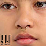 Close-up of a person's face featuring brown eyes, pink lips, and the TIKA Teardrop Septum Clicker Ring in Gold | 16 gauge by Kaya Movement that exemplifies minimalist design. The text "Kaya Movement" is visible in the bottom left corner of the image.
