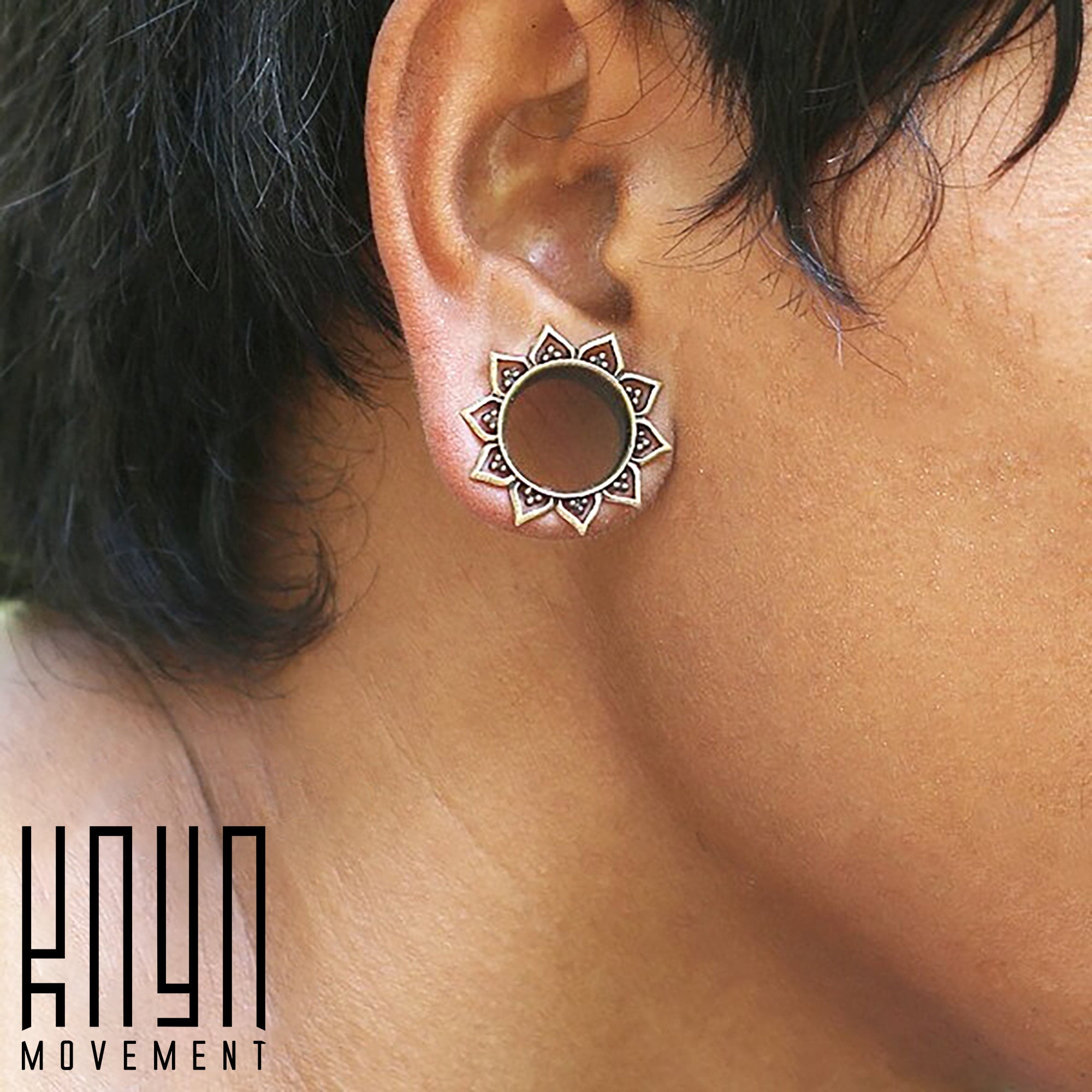 Close-up of a person wearing a circular, floral-patterned ear gauge, featuring a mandala lotus flower design. The silver-colored tunnel is intricately detailed. The image has the text “Kaya Movement” in the bottom left corner and showcases the NIN Sacred Lotus Flower Round Tunnels in Silver, available in sizes ranging from 3mm to 18mm gauge.