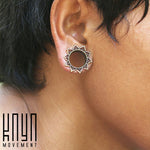 Close-up of a person wearing a circular, floral-patterned ear gauge, featuring a mandala lotus flower design. The silver-colored tunnel is intricately detailed. The image has the text “Kaya Movement” in the bottom left corner and showcases the NIN Sacred Lotus Flower Round Tunnels in Silver, available in sizes ranging from 3mm to 18mm gauge.