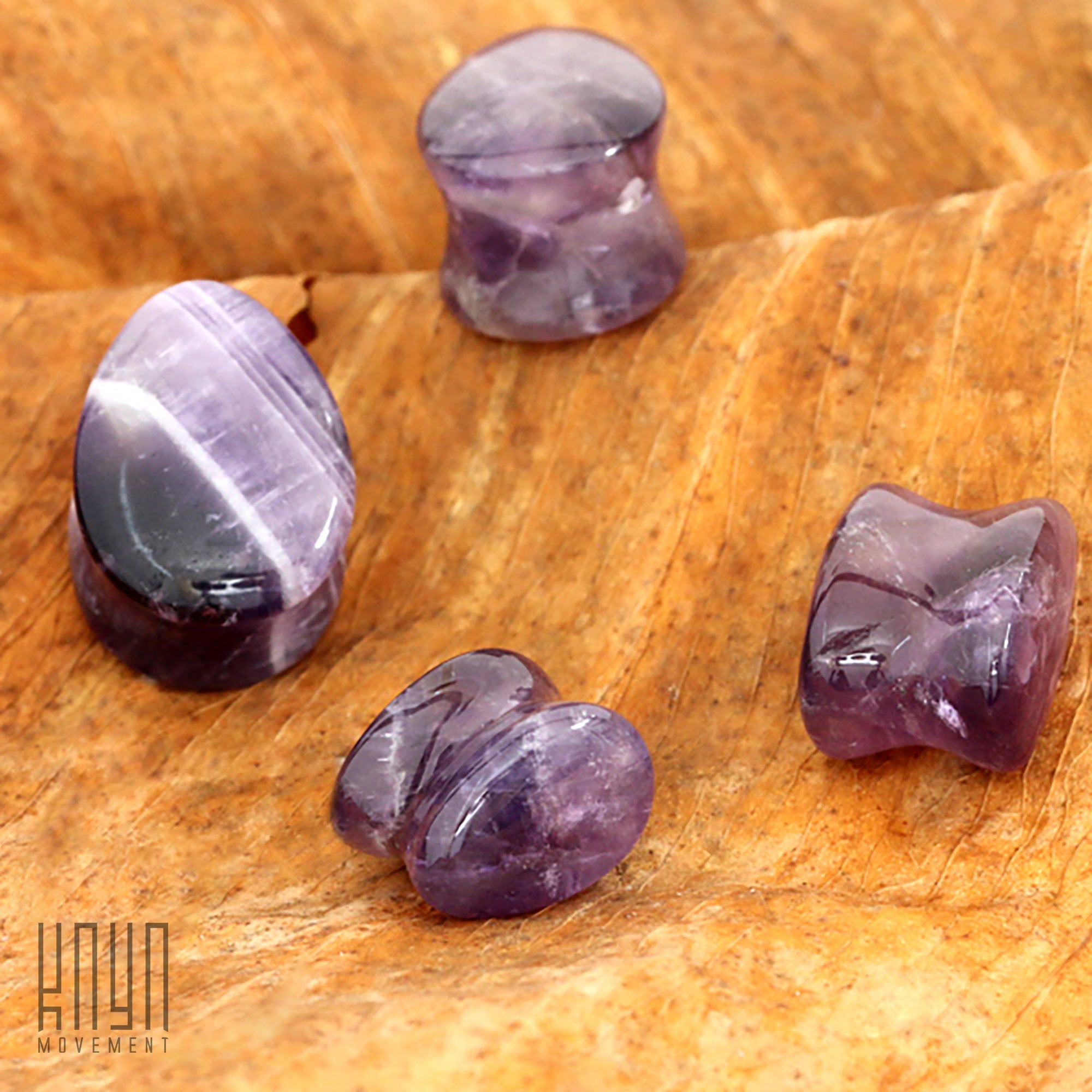 Four polished amethyst teardrop stone saddle plugs of varying sizes, ranging from 8mm to 25mm gauge, are placed on a wooden surface. The stones display natural white streaks and patterns. On the left side of the image is a small logo that reads "Kaya Movement.