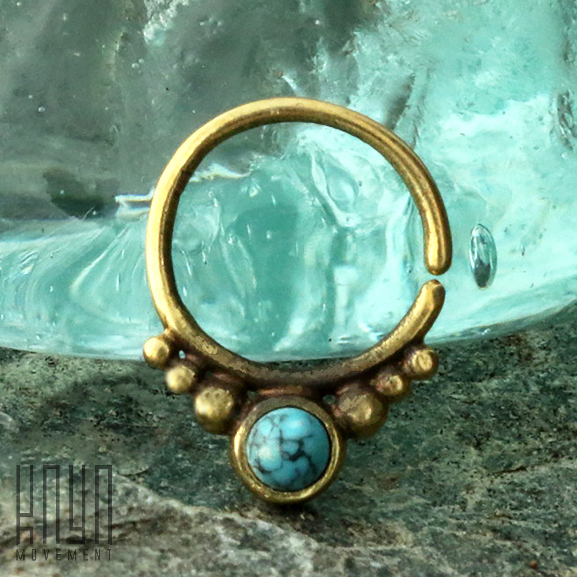 A close-up of the ANI Seamless Septum Ring in Gold & Turquoise by Kaya Movement, showcasing its turquoise gemstone set within the bottom part of the 16 gauge ring. Clusters of small brass balls surround the blue stone, while it is elegantly displayed against a glass and stone background, highlighting its gold brass body jewelry beauty.