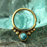 A close-up of the ANI Seamless Septum Ring in Gold & Turquoise by Kaya Movement, showcasing its turquoise gemstone set within the bottom part of the 16 gauge ring. Clusters of small brass balls surround the blue stone, while it is elegantly displayed against a glass and stone background, highlighting its gold brass body jewelry beauty.