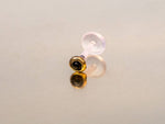 A photograph showcasing the KEA Labret Stud in Gold & Onyx by Kaya Movement, featuring a minimalist design with a gold-colored setting and Black Onyx stone. The 16-gauge earring includes a translucent plastic post and is positioned on a reflective surface, creating a shadow.
