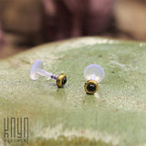 A pair of KEA Labret Studs in Gold & Onyx (16 gauge) lies on a smooth, green, shiny surface. Beside them, the words "Kaya Movement" are subtly visible in the bottom left corner. The background is blurred, emphasizing the minimalist stud earrings crafted with Black Onyx stone jewelry in the foreground.