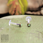 Close-up of two KEA Labret Studs in gold, each featuring a white opal gemstone, resting on a textured green surface. In the background, out-of-focus yellow and green plants add a natural element. The text "Kaya Movement" is visible in the bottom left corner, embodying a minimalist setting with gold brass body jewelry.