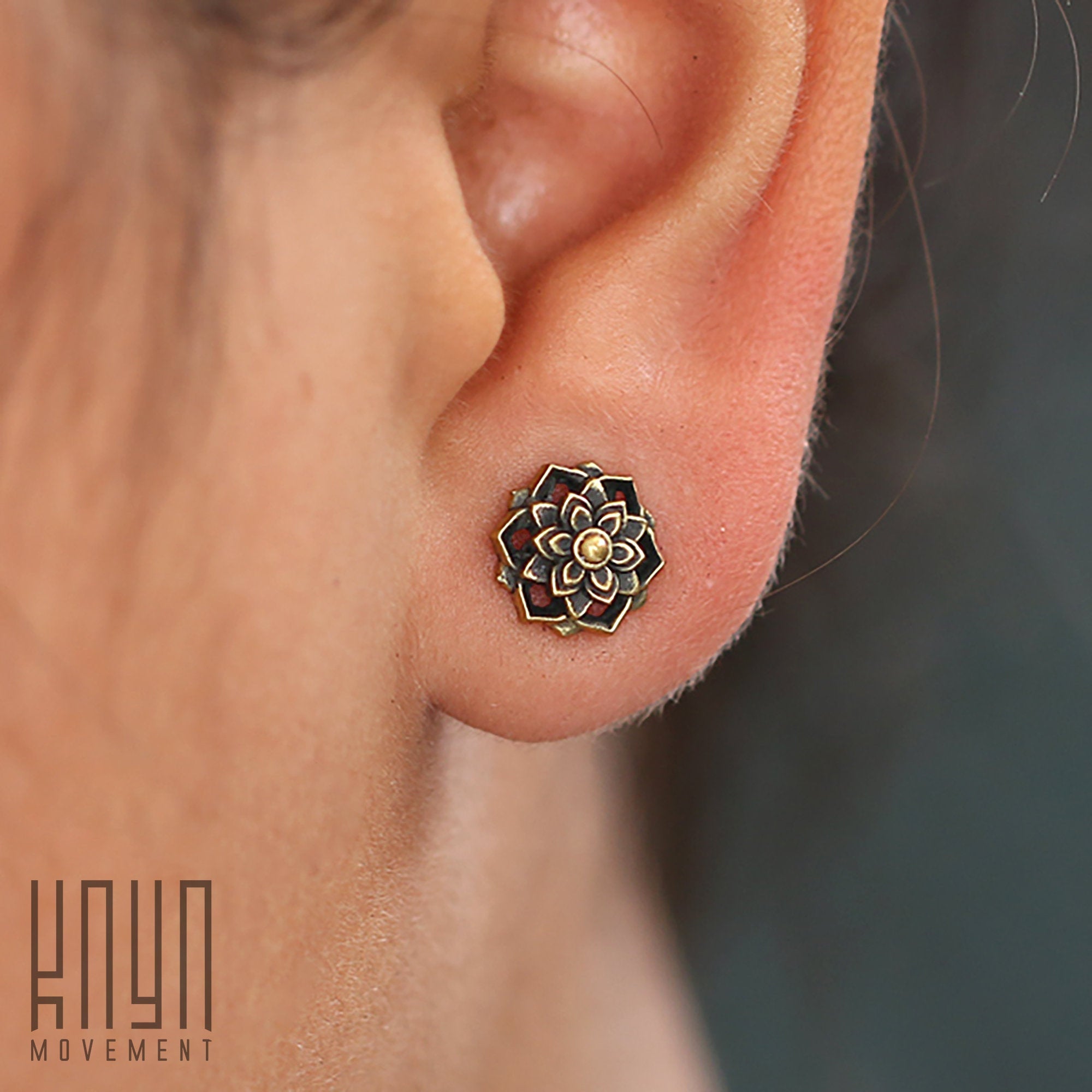 Close-up of an ear lobe showcasing a detailed, floral-patterned ILLY Lotus Mandala stud earring made from yellow brass with an intricate design. The background is blurred, highlighting the earring's craftsmanship. A logo in the bottom-left corner reads "Kaya Movement.