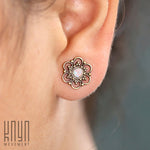 Close-up of an ear adorned with Kaya Movement's Opal Studs Earrings, featuring intricate brass metalwork with tiny bead accents and a central opalescent stone. The image background is blurred, with the "Kaya Movement" logo visible in the bottom left corner.