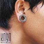 A close-up of a person's ear adorned with the LOTUS Sacred Lotus Mandala Round Tunnel in silver, showcasing its intricate designs. The gauge, reminiscent of a handcrafted tribal plug, is part of Kaya Movement's collection. The bottom left corner features the text "Kaya Movement" in a stylized font.