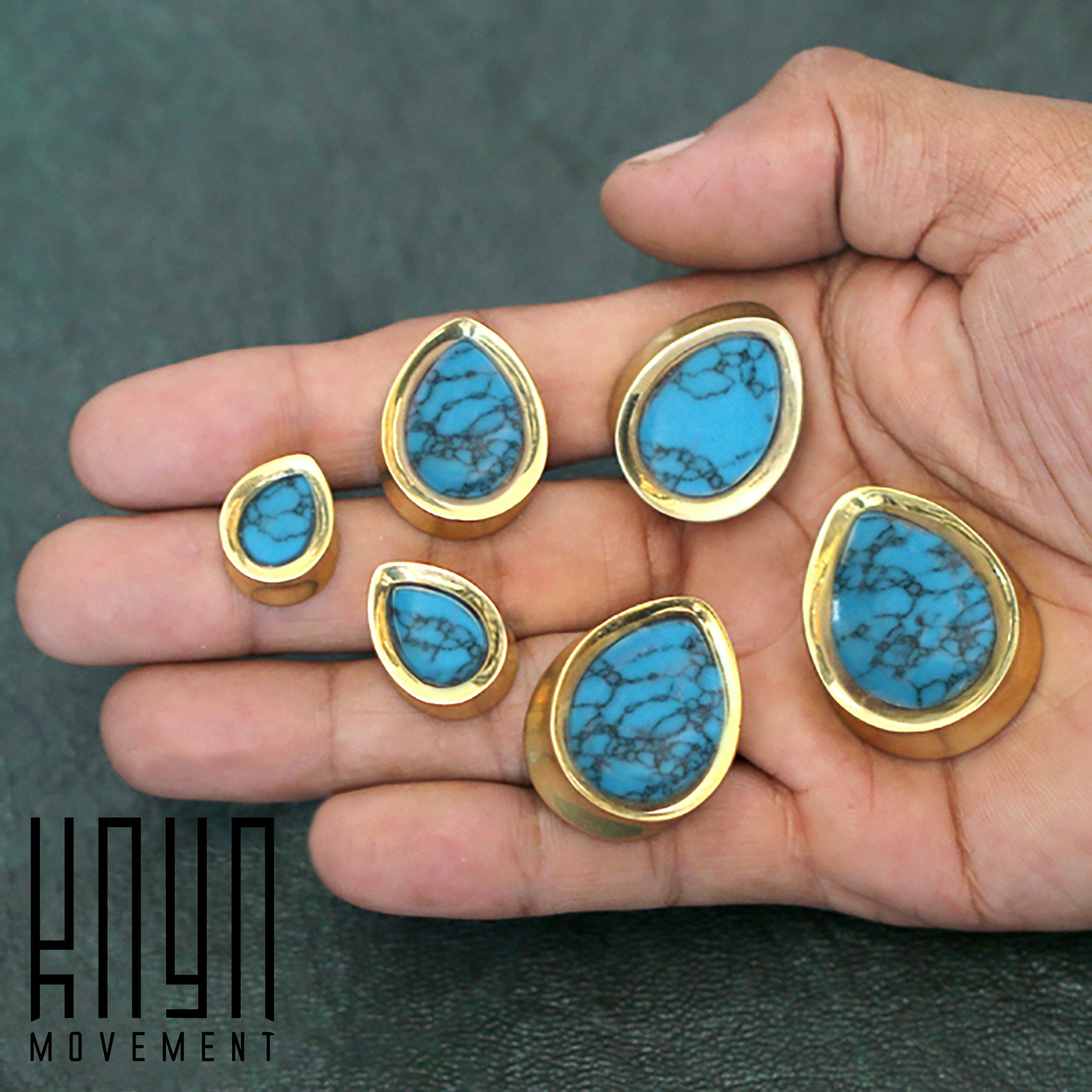 A hand showcases six Teardrop Stone Saddle Plugs in Gold & Turquoise, available in gauges ranging from 12mm to 25mm. These intricate ear jewelry pieces feature delicate veining and double-flared plugs. The text "Kaya Movement" is visible in the bottom left corner of the image.