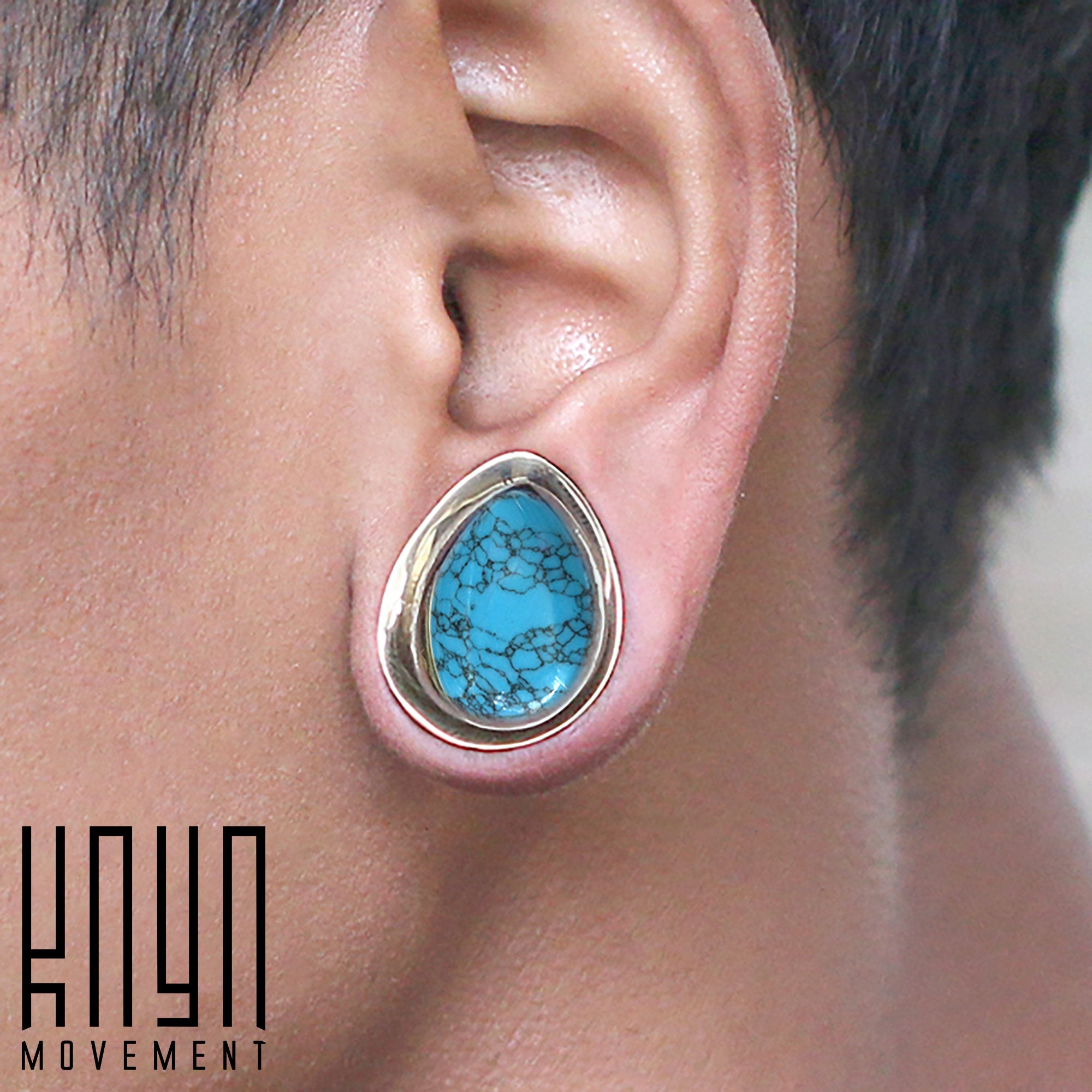 Close-up of a person's ear adorned with a stylish teardrop-shaped earring featuring a turquoise stone inlay framed in gold. The earring showcases intricate patterns on the stone. The logo "Kaya Movement" appears in the bottom left corner, reminiscent of Teardrop Stone Saddle Plugs in Gold & Turquoise from 12mm to 25mm gauge.