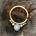 A close-up shot of the ANI Seamless Septum Ring in Gold & Moonstone | 16 gauge, with a moonstone centerpiece surrounded by small gold beads. The exquisite piece of body jewelry is resting on a textured wooden surface, with the Kaya Movement logo visible in the lower left corner.