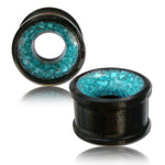 Two TOAN Round Saddle Tunnels in Wood & Turquoise by Kaya Movement are shown. The turquoise inlays have a marbled, textured appearance. One gauge is standing upright, displaying the detailed inlay, while the other lies flat, highlighting its circular shape. These gauges resemble organic tunnels carved from Ironwood and come in sizes ranging from 4mm to 25mm.