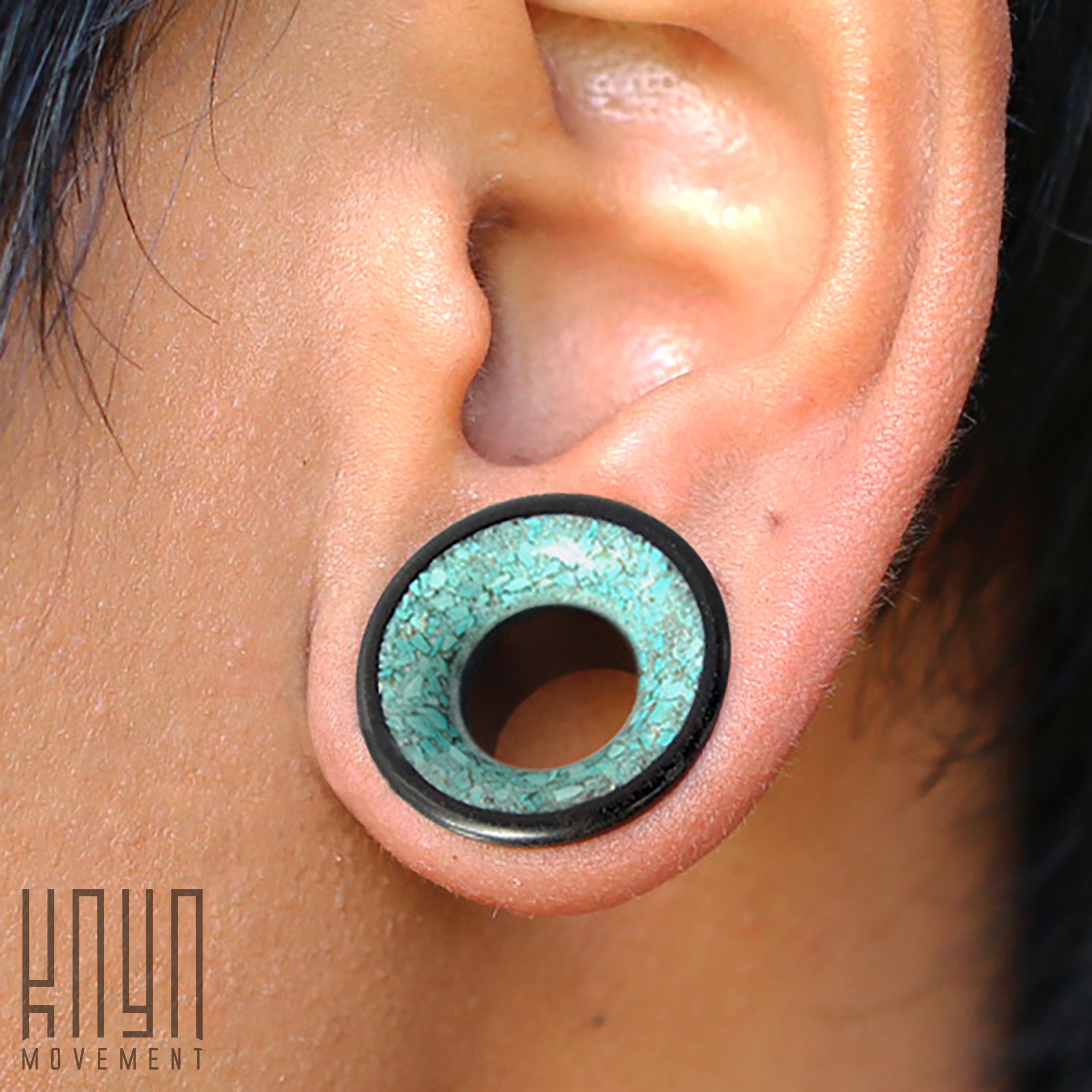 Close-up image of a person's ear with a stretched earlobe featuring a large gauge plug. The TOAN Round Saddle Tunnel by Kaya Movement displays an ironwood construction with a black outer rim and a turquoise inlay center that offers a textured appearance. Available in sizes ranging from 4mm to 25mm.