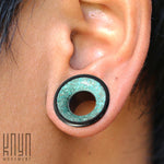 Close-up image of a person's ear with a stretched earlobe featuring a large gauge plug. The TOAN Round Saddle Tunnel by Kaya Movement displays an ironwood construction with a black outer rim and a turquoise inlay center that offers a textured appearance. Available in sizes ranging from 4mm to 25mm.