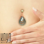 A close-up image showcases a person's belly button adorned with the exquisite IBOSA Teardrop Belly Piercing Ring in Gold & Abalone Shell by Kaya Movement. The jewelry features a gold-colored ball piercing above the navel and an ornate, teardrop-shaped piece hanging below. This teardrop, showcasing intricate designs and an abalone shell centerpiece, is accentuated by a gently placed hand on the skin to the right.