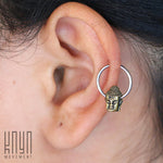 Close-up of a person's ear showcasing the BUDDHA Silver Ring with a gold Buddha head charm from Kaya Movement, worn in the helix part of the ear. The logo "Kaya Movement" is visible in the bottom left corner.