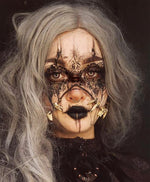 A person with long, wavy, gray hair is wearing elaborate black makeup that resembles a mask with intricate designs. They have minimalist Kaya Movement MOON Seamless Rings in Gold (16 gauge) around their mouth and nose. Wearing dark clothing and displaying a serious expression, their black lipstick accentuates the look perfectly.