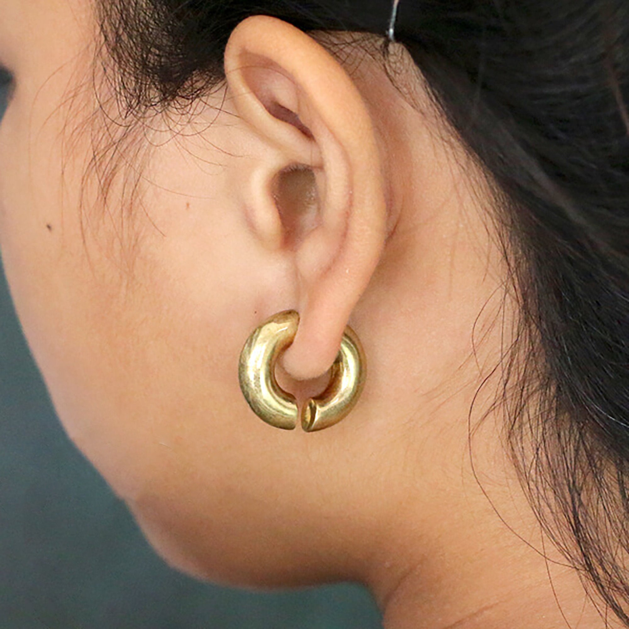 Close-up of a person’s ear wearing a chunky gold hoop earring from Kaya Movement. The ELI Fake Hoop Gauge Earrings in Gold, designed to resemble fake gauge earrings, have a smooth texture. The person's hair is pulled back, revealing the 18 gauge golden jewelry with its 925 silver post clearly. The background is blurred.