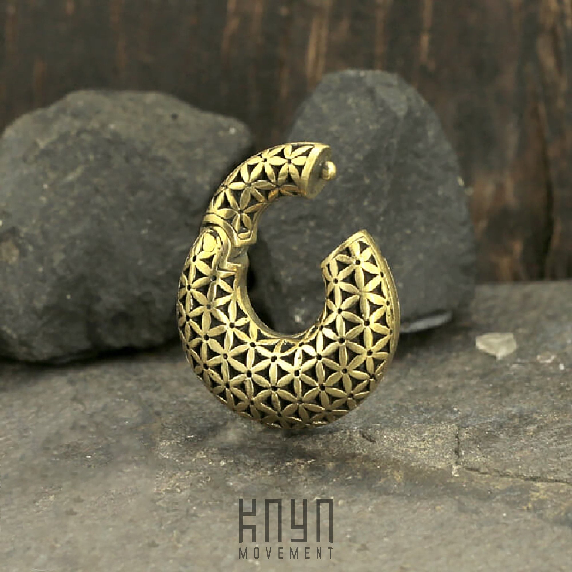 The FLOWER OF LIFE Hoop Ear Weights in Gold with Clicker by Kaya Movement, designed in a striking crescent moon shape, stand upright against a backdrop of dark, textured rocks. The ear weights' geometric design features interlocking triangles and a flower of life pattern, creating a visually intriguing contrast with their natural stone surroundings.