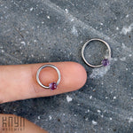A hand showcases the ORASA Nose Ring in Silver & Amethyst (16 gauge) from Kaya Movement, elegantly balanced on the index finger. Another identical nose ring rests on a gray, speckled surface nearby. The text "Kaya Movement" is visible in the bottom left corner.