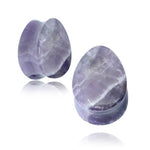 Two polished, teardrop-shaped stones with a light purple hue and translucent appearance. The stones have subtle white veining and are displayed against a white background, with a soft reflection visible beneath them. These Amethyst Teardrop Stone Saddle Plugs by Kaya Movement are perfect for spiritual enhancement, available in gauges ranging from 8mm to 25mm.