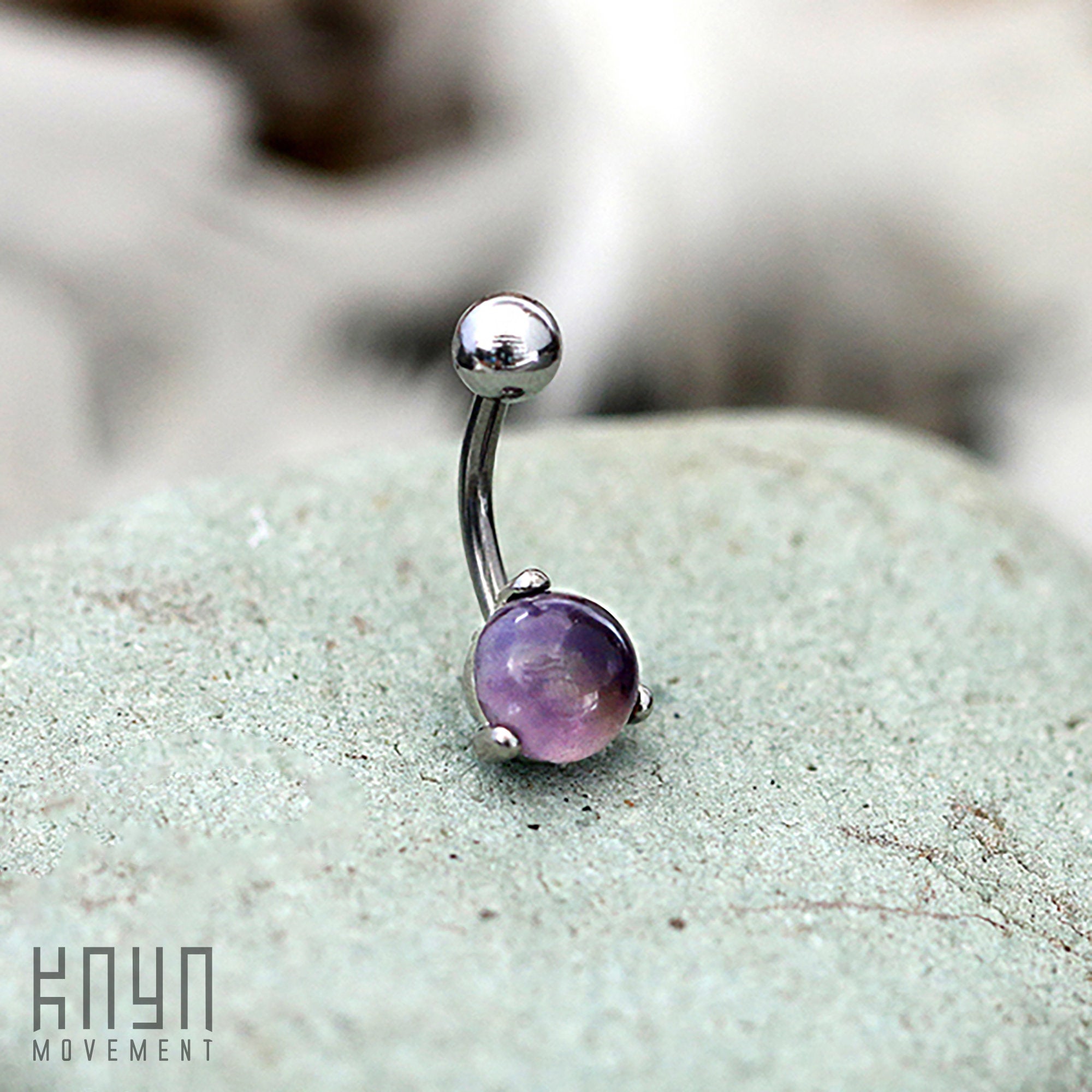 A 14-gauge Amethyst Belly Piercing Ring in Silver from Kaya Movement, crafted from 316L surgical steel and featuring a natural amethyst stone at one end, is placed on a gray stone surface. "Kaya Movement" is inscribed in the bottom left corner. The softly blurred background draws attention to this exquisite piece of navel jewelry as the central focus.