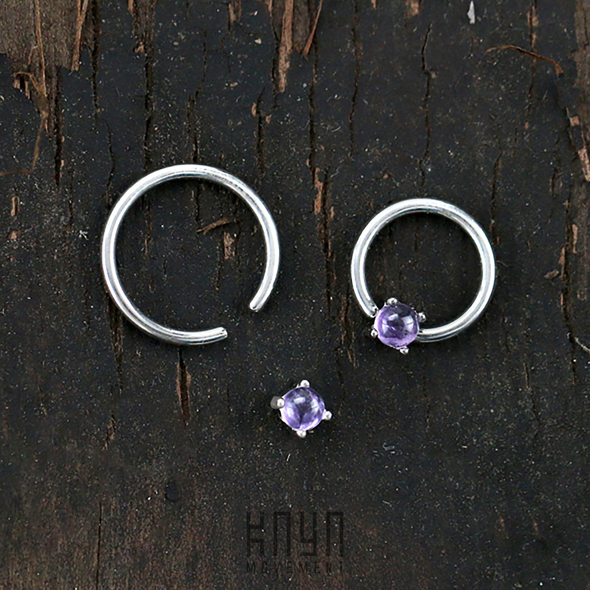 A small circular Kaya Movement ORASA Nose Ring in Silver & Amethyst (16 gauge) along with an open silver nose ring and a matching purple gemstone earring stud are arranged on a dark wooden background. The image, showcasing exquisite silver body jewelry, has a textured, rustic feel.