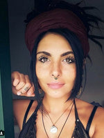 A woman with a Kaya Movement SYN Seamless Septum Ring in Gold & Onyx, along with tattoos on her fingers, poses for a photo. She has dark eyes, long eyelashes, and a beauty mark on her cheek. Her hair is wrapped in a headscarf adorned with gold brass body jewelry, and she wears a necklace featuring a black onyx gemstone. Her sleeveless outfit is visible.