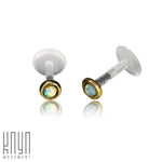 A pair of KEA Labret Studs by Kaya Movement, made of clear plastic with natural White Opal stones set in gold-colored bezels. These opals exhibit an iridescent play of color. The background is white, and the bottom left corner showcases the text "Kaya Movement" in a stylish, modern font.