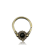 The SYN Seamless Septum Ring in Gold & Onyx by Kaya Movement is a small, circular brass nose ring showcasing an intricate design with a central black onyx gemstone, surrounded by tiny decorative elements. Its open hoop design ensures easy placement and is beautifully displayed against a plain white background.