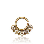 The AIA Seamless Septum Ring in Gold by Kaya Movement is an 18-gauge nose ring showcasing an ornate filigree design with heart-shaped patterns and small decorative elements along the bottom edge. Enhanced with a subtle tribal ethnic flower motif, this ring features an open segment for easy insertion. Its intricate, symmetrical layout is polished to perfection.