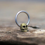 The SKULL Gothic Silver Ring with Gold Pendant by Kaya Movement is artfully displayed on a textured surface. The ring, available in 18, 16, or 14 gauge, features a striking gold-colored skull pendant at the base of a sleek silver-colored band. The focus on the ring is sharp, while the blurred background lends a rustic feel to the image.
