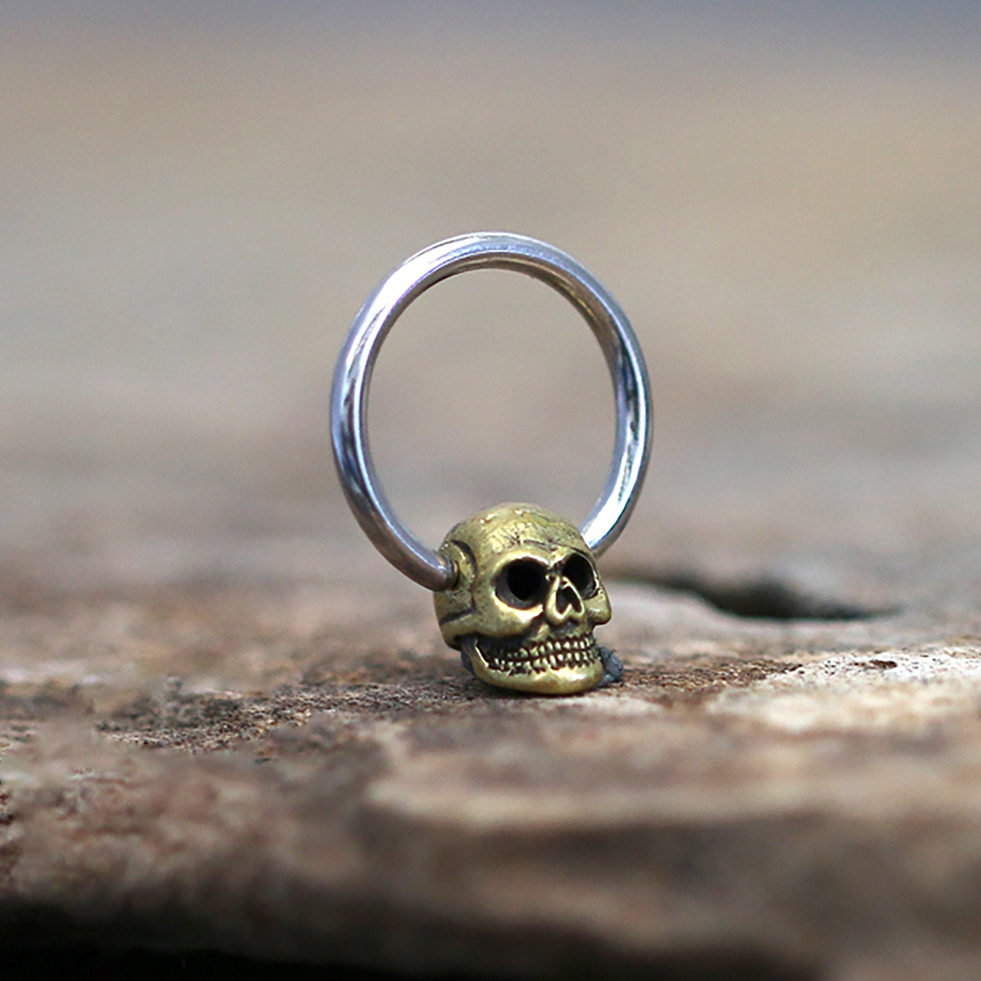 The SKULL Gothic Silver Ring with Gold Pendant by Kaya Movement is artfully displayed on a textured surface. The ring, available in 18, 16, or 14 gauge, features a striking gold-colored skull pendant at the base of a sleek silver-colored band. The focus on the ring is sharp, while the blurred background lends a rustic feel to the image.