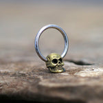 The SKULL Gothic Silver Ring with Gold Pendant by Kaya Movement is artfully displayed on a textured surface. The ring, available in 18, 16, or 14 gauge, features a striking gold-colored skull pendant at the base of a sleek silver-colored band. The focus on the ring is sharp, while the blurred background lends a rustic feel to the image.